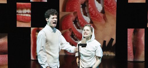 Auf der Bühne stehen eine Frau und ein Mann vor einem Mikrofon. Im Hintergrund sind Lippen übergroß projiziert.