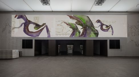 Large wall installation with abstract colored shapes above a black rectangular platform in an exhibition space.