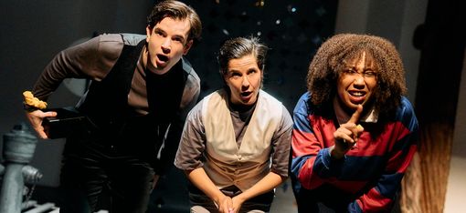 Three people are standing close together: two of them look on in amazement, whilst the third person is smiling and holding up their index finger.