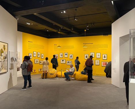 People viewing artworks on yellow walls in a museum exhibition with seating cushions in the center.