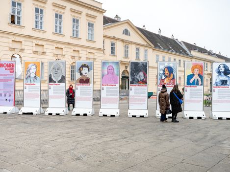 nine large information pillars with portrait photos and texts about women as part of  the exhibition  „women against war“; three women are looking at the pillars in front of the MQ.