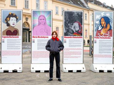 Four large poster stands with stylised portraits of women and texts are located outdoors in front of the Museumsquartier Wien, with a person standing in front of them.