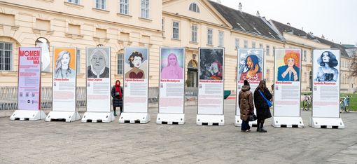 nine large information pillars with portrait photos and texts about women as part of  the exhibition  „women against war“; three women are looking at the pillars in front of the MQ.
