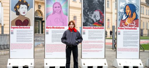 Four large poster stands with stylised portraits of women and texts are located outdoors in front of the Museumsquartier Wien, with a person standing in front of them.