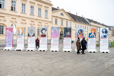 nine large information pillars with portrait photos and texts about women as part of  the exhibition  „women against war“; three women are looking at the pillars in front of the MQ.
