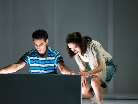 Two young people look at a glowing surface, one sitting wearing a striped shirt, the other standing wearing a light dress with a jacket.