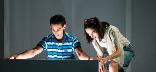 Two young people look at a glowing surface, one sitting wearing a striped shirt, the other standing wearing a light dress with a jacket.
