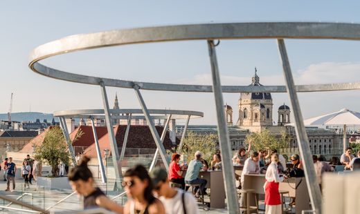 Menschen sitzen und stehen auf einer modernen Dachterrasse mit Metallringen und Glasgeländern. Einige unterhalten sich an Stehtischen unter Sonnenschirmen. Im Hintergrund sind historische Gebäude mit Kuppel und Türmen zu sehen, die über die Dächer der Stadt hinausragen.