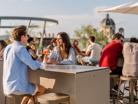 Zwei Personen sitzen an einem Tisch auf der MQ Libelle und trinken Getränke, während sie sich unterhalten, im Hintergrund weitere Gäste und eine Kuppel eines historischen Gebäudes