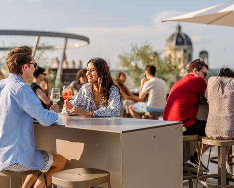 Zwei Personen sitzen an einem Tisch auf der MQ Libelle und trinken Getränke, während sie sich unterhalten, im Hintergrund weitere Gäste und eine Kuppel eines historischen Gebäudes