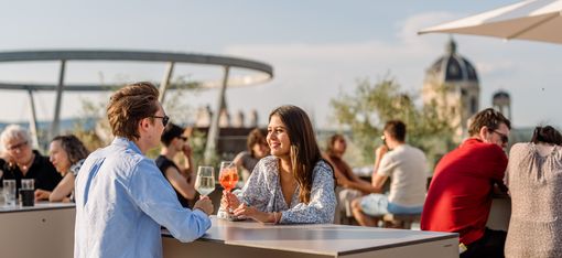 Zwei Personen sitzen an einem Tisch auf der MQ Libelle und trinken Getränke, während sie sich unterhalten, im Hintergrund weitere Gäste und eine Kuppel eines historischen Gebäudes