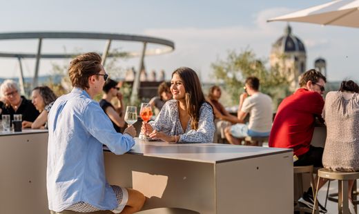 Two people are sitting at a table on the MQ Libelle, drinking beverages while chatting, with other guests and the dome of a historic building in the background.