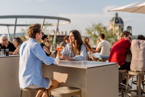 Zwei Personen sitzen an einem Tisch auf der MQ Libelle und trinken Getränke, während sie sich unterhalten, im Hintergrund weitere Gäste und eine Kuppel eines historischen Gebäudes