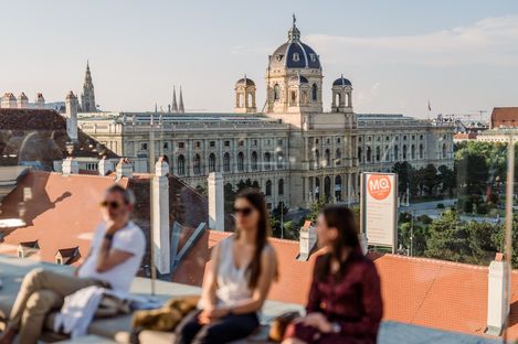 Besucher:innen sitzen auf der Dachterrasse Zur Libelle im MuseumsQuartier Wien, im Hintergrund das Kunsthistorische Museum und die Wiener Innenstadt.