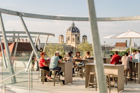 Menschen sitzen an Tischen auf der MQ Libelle. Im Hintergrund die Dächer Wiens.