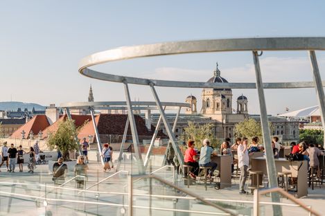 Two people clink drinks on the rooftop terrace Zur Libelle at MuseumsQuartier Vienna, with the city of Vienna in the background.