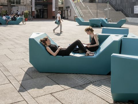 Zwei Personen entspannen auf türkisfarbenen Enzi-Sitzmöbeln im sonnigen Innenhof des MuseumsQuartier Wien, im Hintergrund das mumok und weitere Besucher:innen.