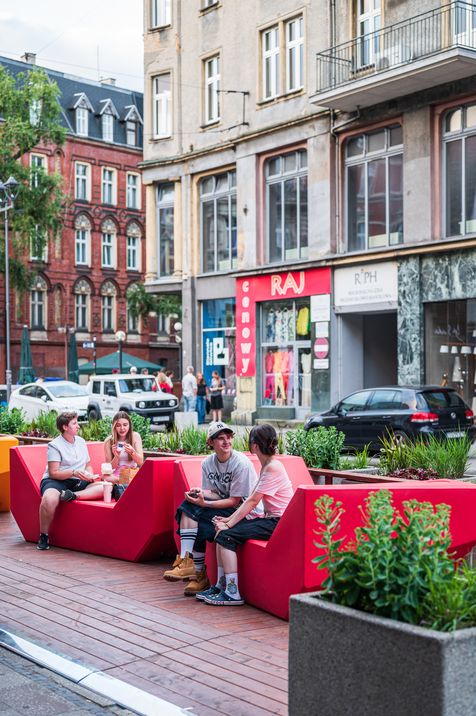 Vier junge Menschen sitzen auf roten Enzi-Sitzmöbeln in einem urbanen Straßenraum und unterhalten sich, umgeben von Pflanzbeeten und historischen Gebäuden.