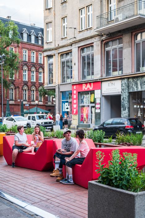 Vier junge Menschen sitzen auf roten Enzi-Sitzmöbeln in einem urbanen Straßenraum und unterhalten sich, umgeben von Pflanzbeeten und historischen Gebäuden.