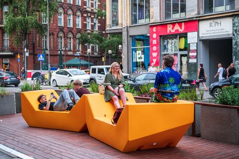 Mehrere Personen nutzen gelbe Enzi-Sitzmöbel zum Sitzen und Liegen in einem belebten Stadtraum mit Geschäften, Autos und Fußgänger:innen.