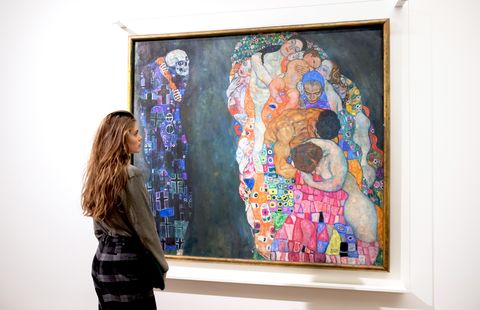 A woman stands thoughtfully in front of a colorful painting by Gustav Klimt featuring intertwined human figures and a skull, evoking themes of life and death.