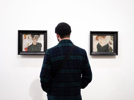 Person von hinten vor zwei gerahmten Porträtzeichnungen an weißer Wand
