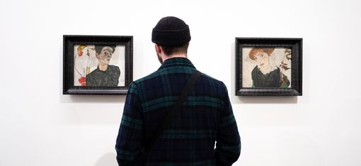 Person von hinten vor zwei gerahmten Porträtzeichnungen an weißer Wand