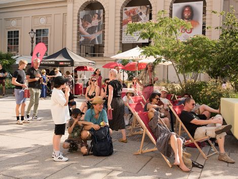 Menschen sitzen und stehen in einem sonnigen Innenhof mit Liegestühlen, Sonnenschirmen und einem Infostand vor einem historischen Gebäude.