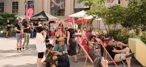 Menschen sitzen und stehen in einem sonnigen Innenhof mit Liegestühlen, Sonnenschirmen und einem Infostand vor einem historischen Gebäude.