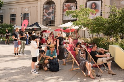 Menschen sitzen und stehen in einem sonnigen Innenhof mit Liegestühlen, Sonnenschirmen und einem Infostand vor einem historischen Gebäude.