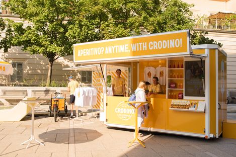 Gelb-weißer Verkaufsstand mit der Aufschrift 'APERITIVO? ANYTIME, WITH CRODINO!' und mehreren Personen in gelb-weiß gestreiften Shirts in einem sonnigen Außenbereich mit Bäumen.