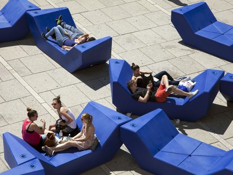 Menschen entspannen auf blauen Enzi-Sitzmöbeln im MuseumsQuartier Wien, einige liegen oder sitzen in kleinen Gruppen auf dem gepflasterten Platz.