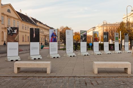 Several upright information panels with images and texts on a paved square between two stone benches and historic buildings in the background