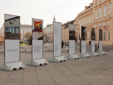 Five vertical posters on concrete stands displaying photos and texts on a paved square in front of historic buildings.