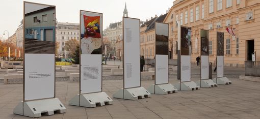 Five vertical posters on concrete stands displaying photos and texts on a paved square in front of historic buildings.