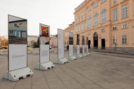 Several freestanding information panels with text and images are arranged on a paved square in front of a historic building with large windows.