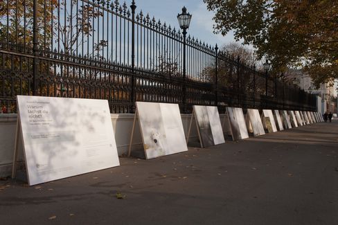 Several large posters are lined up along a sidewalk in front of a tall wrought-iron fence with lanterns, with trees showing autumn foliage in the background.