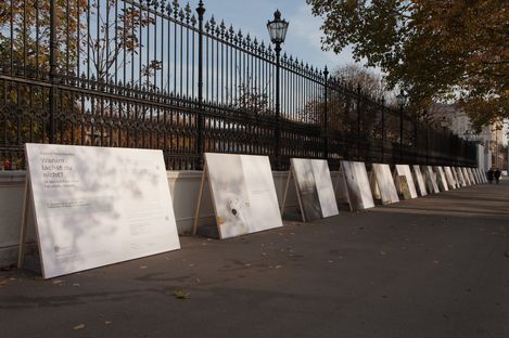 Several large posters are lined up along a sidewalk in front of a tall wrought-iron fence with lanterns, with trees showing autumn foliage in the background.