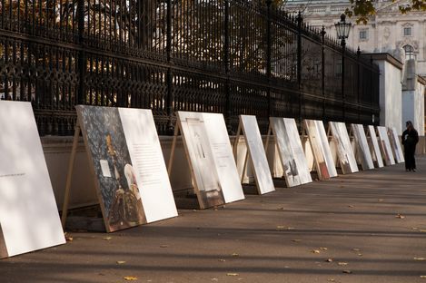 Several large panels with images and text leaning against an iron fence along a sidewalk with a person standing at the right edge.