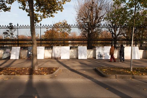 Two people walk along a street past a row of large white panels leaning against a fence surrounded by trees with autumn leaves.