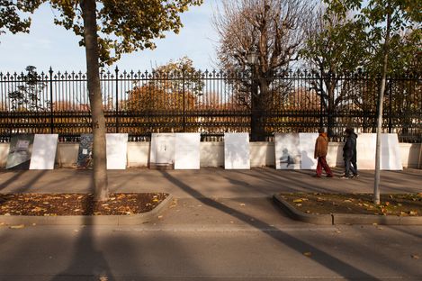 Two people walk along a street past a row of large white panels leaning against a fence surrounded by trees with autumn leaves.