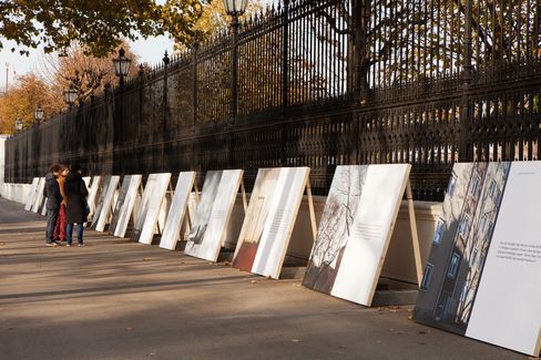 Several large posters standing on the ground along a wrought-iron fence, two people viewing the display.