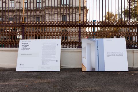 Two large posters leaning against an ornate metal fence in front of a historic building, one with text and one with a photo of white walls and a window frame.