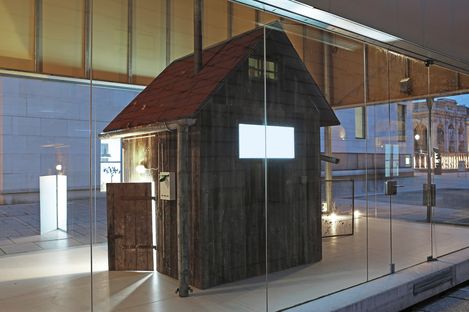 Installation of a small wooden house in the MQ Art Box at night, with illuminated interior and open entrance.