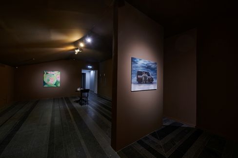 Dimly lit exhibition room with two paintings on the walls and a table with a chair in the center