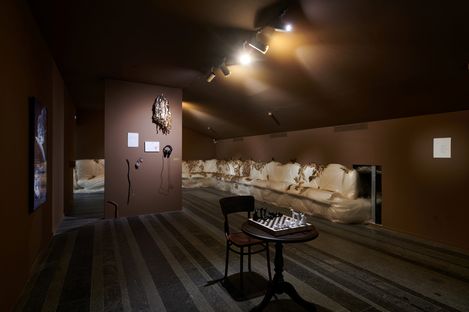 Dimly lit room with brown walls, a long white bench along the right wall, a small round table with a chessboard and two chairs in the center, and artworks on the walls.