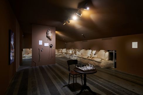 Dimly lit room with brown walls, a long white bench along the right wall, a small round table with a chessboard and two chairs in the center, and artworks on the walls.