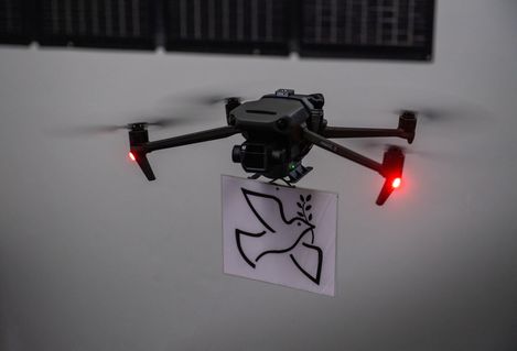 Black drone with red lights carrying a sheet showing a stylized dove with an olive branch against a white wall