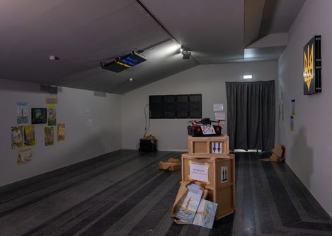 Dimly lit room with several wooden crates, posters on the wall, and an illuminated picture with a yellow symbol