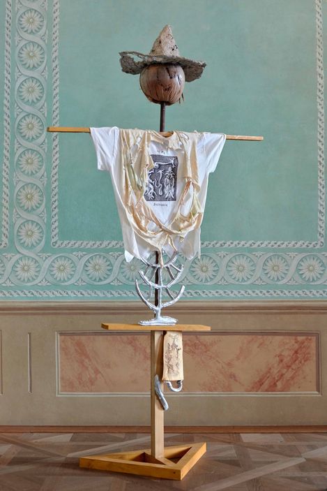 Wooden sculpture wearing a straw hat, white T-shirt with black design, and hanging fabric, standing against a light green wall with white decorative pattern.